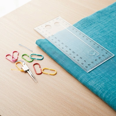 An array of colorful stitch markers and a ruler laid out on a crafting table next to a piece of fabric, ready for use to maintain even stitch spacing.